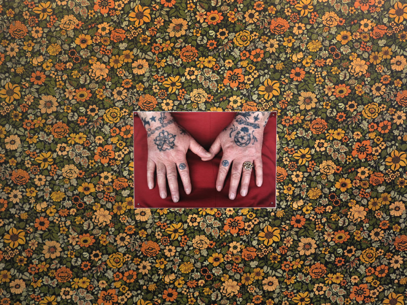 Blumentapete mit einem Foto im Mittelpunkt. Darauf sind alte tattoowierte Hände zu sehen.