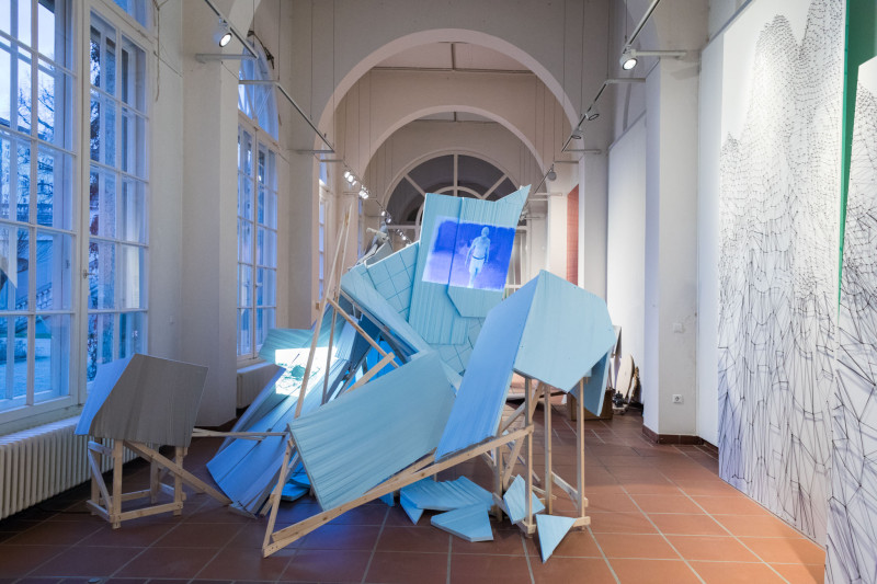 In the middle of the gallery space is an expansive scaffolding consisting of wooden beams and panels fixed to them. A video is projected onto the blue panels.