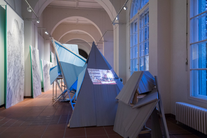 In the middle of the gallery space is an expansive scaffolding consisting of wooden beams and panels fixed to them. A video is projected onto the blue panels.