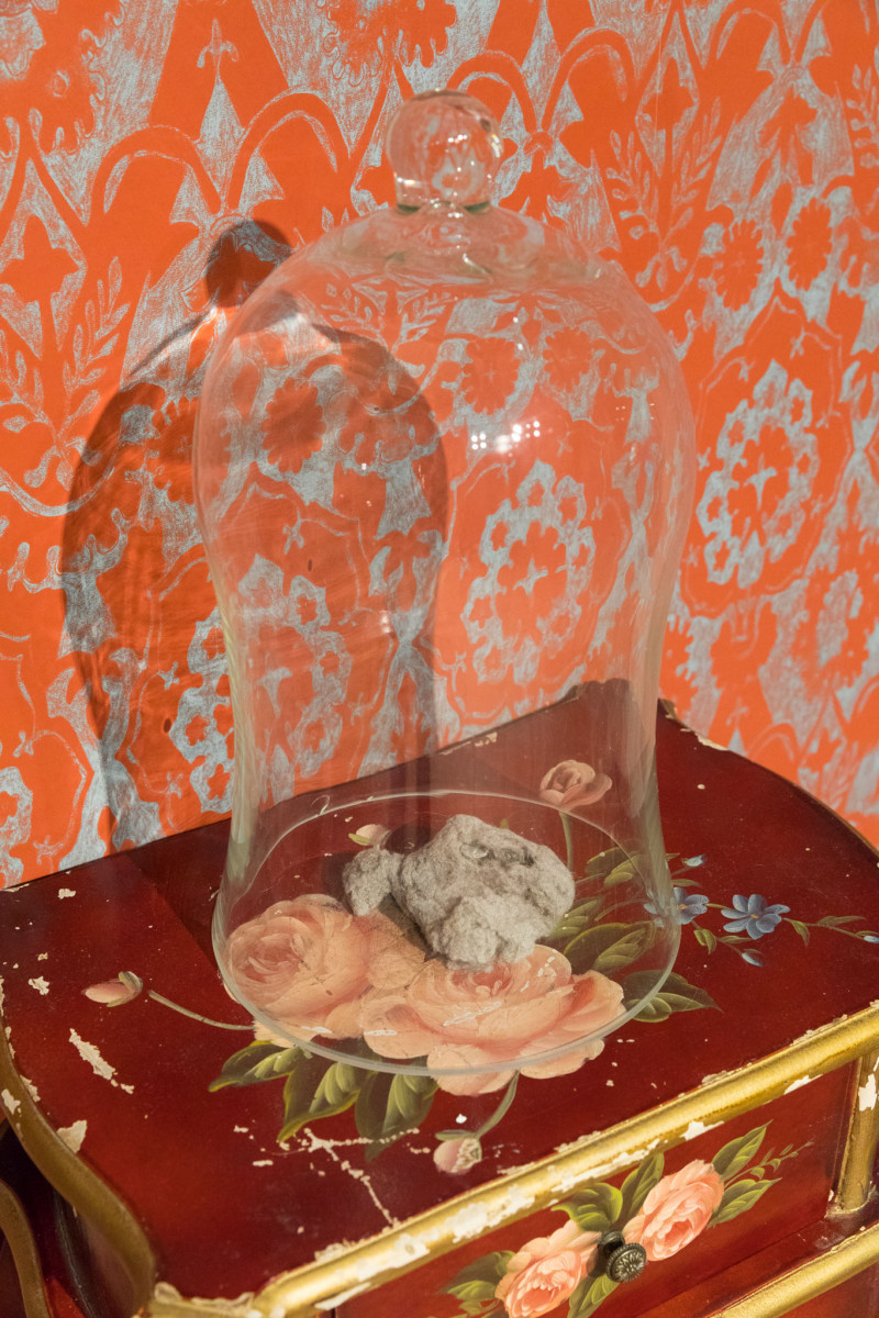 Glass bonnet on a flower table with dust in front of a patterned wallpaper.