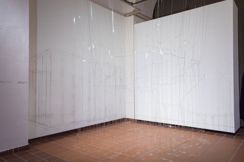 Room installtion. A net of transparent plastic threads hangs down from the ceiling. A cage of delicate lines is drawn on the wall behind.