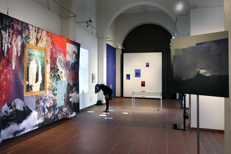 Eine Sicht in den Ausstellungsraum: rechts ein Schild mit der Ansicht eines Bergs, rechts ein großes buntes Textil, dahinter eine Besucherin, die sich über Arbeiten auf dem Boden beugt.