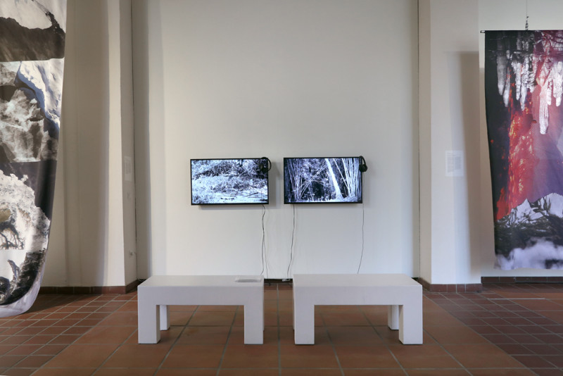 Im Vordergrund stehen zwei weiße Bänke, im Hintergrund sind zwei Monitore an einer Wand angebracht.