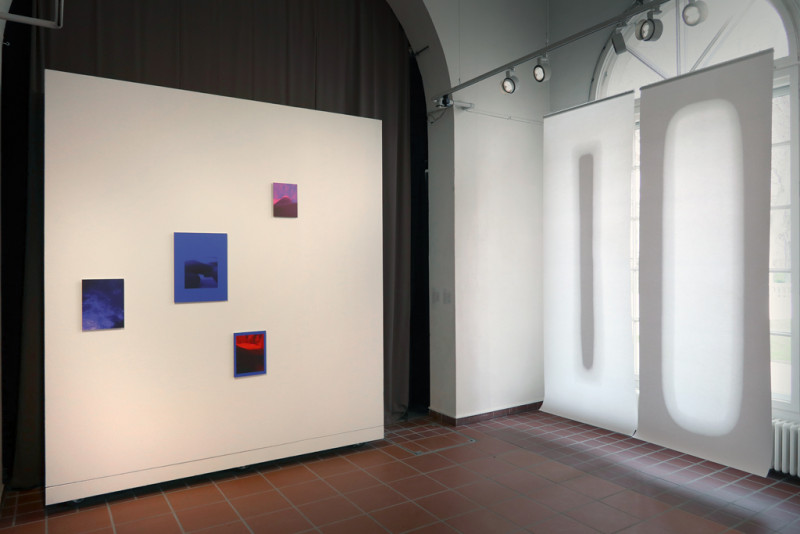 Eine schräge Raumansicht zeigt links eine Wand mit vereinzelten blauen Fotografien, echts zwei vor den Fenstern hängenden Papierstreifen in unterschiedlichen weiß Schatierungen.