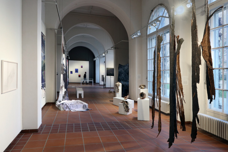Eine Sicht in den Raum der Galerie des Körnerpark mit von der Decke hängenden Baumrinden im Vordergrund und weiteren Kunstwerken im Raum.