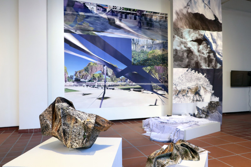 Im Hintergrund ist eine blaue abstrakte Tapete zu sehen sowie ein von der Decke hängendes Textil. Im Vordergrund sind zwei Sockel mit Papierobjekten in Steinobjekt platziert.