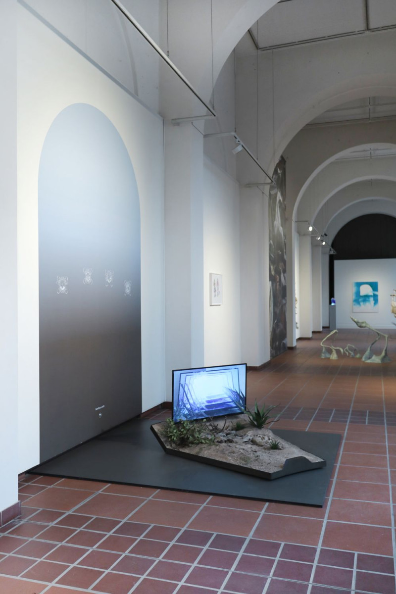 Ein Bildschirm steht auf einem niedrigen, auf dem Boden stehenden Kasten, der mit Sand und Pflanzen gefüllt ist, im Ausstellungsraum.