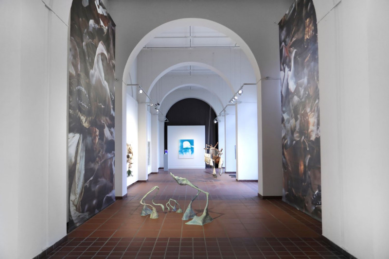 Blick in die Galerie. Auf der linken und rechten Seite hängen große bedruckte Stoffbahnen herunter. Dazwischen sieht man auf dem Boden stehende Skulpturen, die unbekannte Wesen darzustellen scheinen.