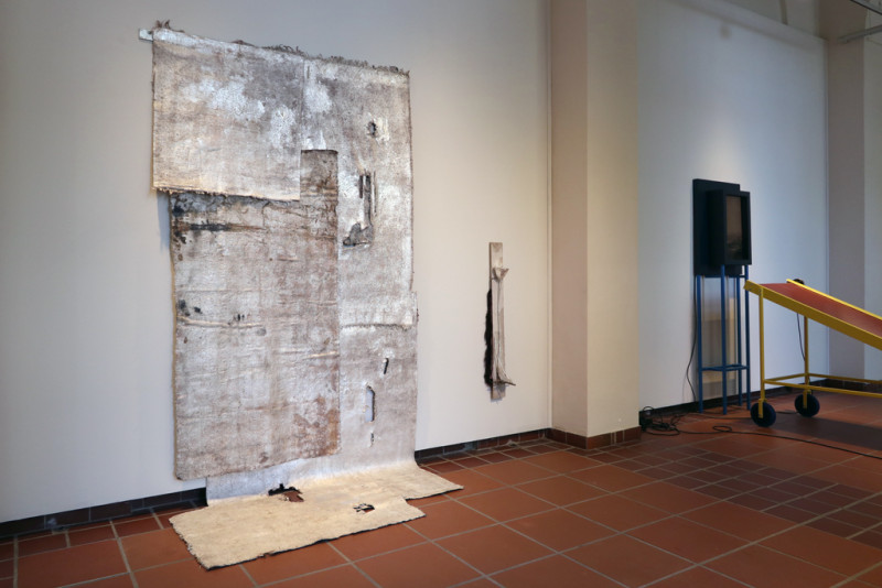 BEin großes aus verschiedenen Teilen zusammengesetztes silbergefärbtes Teppichstück hängt von der Wand und reicht über den Boden.