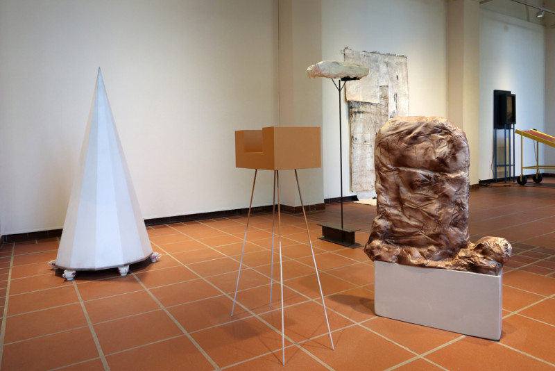 Im Galerieraum sind vier Skulpturen zu sehen, die allesamt auf dem Boden stehen und sehr verschiedene Formen aufweisen, z.B. ein großer Kegel, der auf Teetassen balanciert, oder ein vielkantiger kleinerer Körper, der von vier dünnen, langen Stäben gehalten wird.