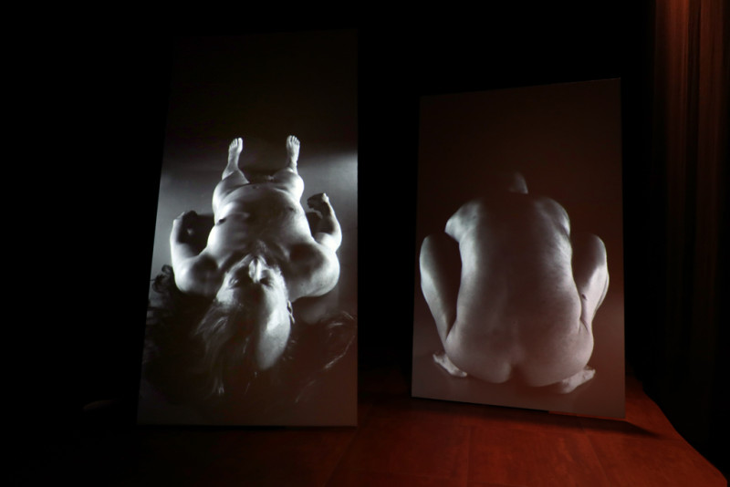 Auf zwei großen nebeneinanderstehenden Monitoren sind lebensgroße nackte menschliche Körper zu sehen. Eine Figur liegt auf dem Boden, die andere ist in einer tiefen Hocke von hinten zu sehen.