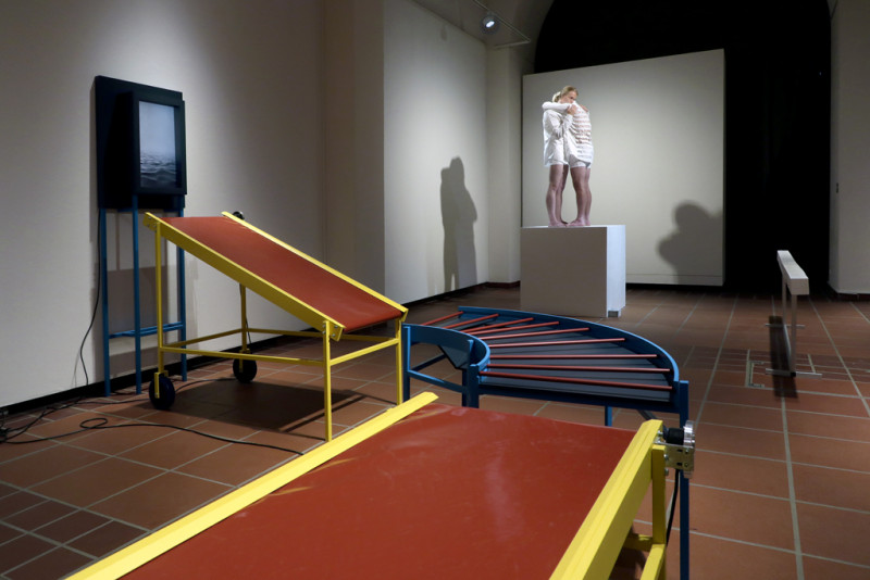 Drei verschiedenfarbige stilisierte Transportbänder sind in einem Bogen hintereinander aufgestellt. Ein Transportband reicht zu einem Bildschirm an der Wand, auf dem Wasseraufnahmen zu sehen sind.