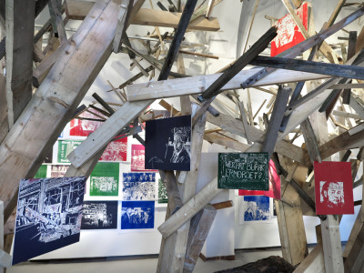 Installation view. A mesh of wooden beams stand in the room and reaches up to the ceiling. Colourful picture prints hang from the beams.