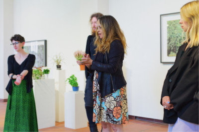 Female curator stands in the exhibition space and speaks while performing a hand gesture. Three people are standing around her. Plants are placed on pedestals behind them