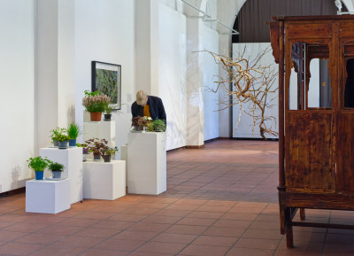 View of the gallery space with a picture on the wall, a wooden bed, an installation of dry branches and plants in flower pots placed on pedestals of different heights. A man bends over one of these plants