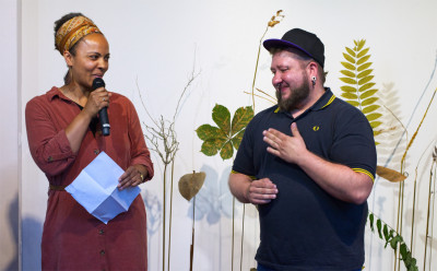 The female curator speaks into a microphone, holds a sheet of paper in her hand and looks over to the sign language interpreter. He holds a hand in front of his chest and makes a gesture