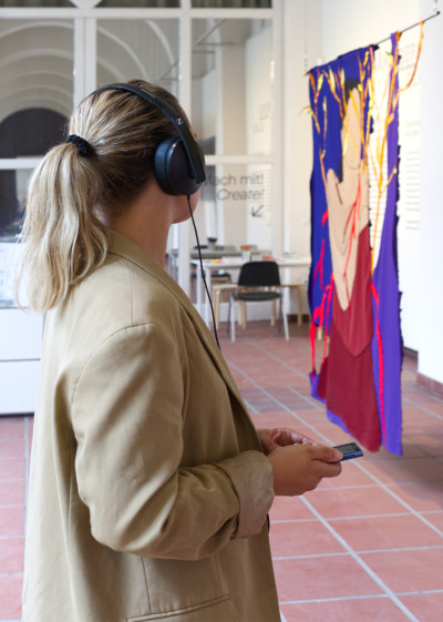 A woman with headphones and an MP3 player in her hand stands in the exhibition space and looks in the direction of an artwork in the form of a hanging fabric