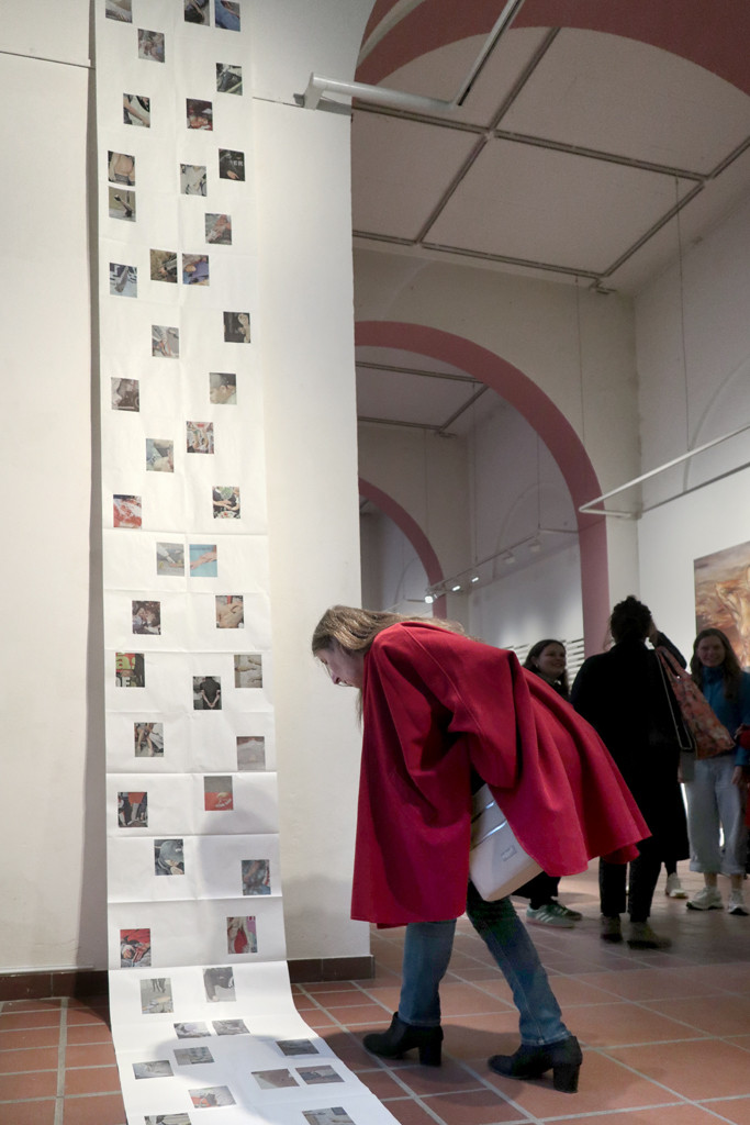 Von der Decke bis auf den Boden der Galerie zieht sich eine Papierbahn, auf die viele kleine Fotografien gedruckt sind. Eine Frau beugt sich herab, um die Fotografien zu betrachten.