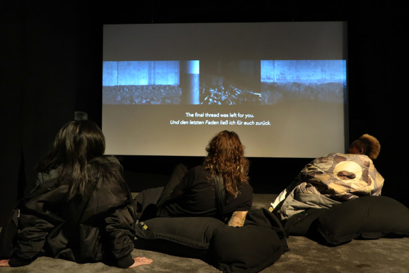 drei Menschen sitzen auf sitzkissen in einem dunklen Raum vor einer Videoinstallation
