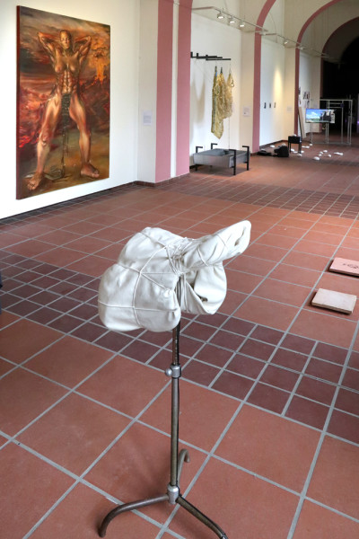 A saddle encompassed by cottonfabric and rope, mounted on a metal stand, the Galerie im Körnerpark can be seen in the backgound