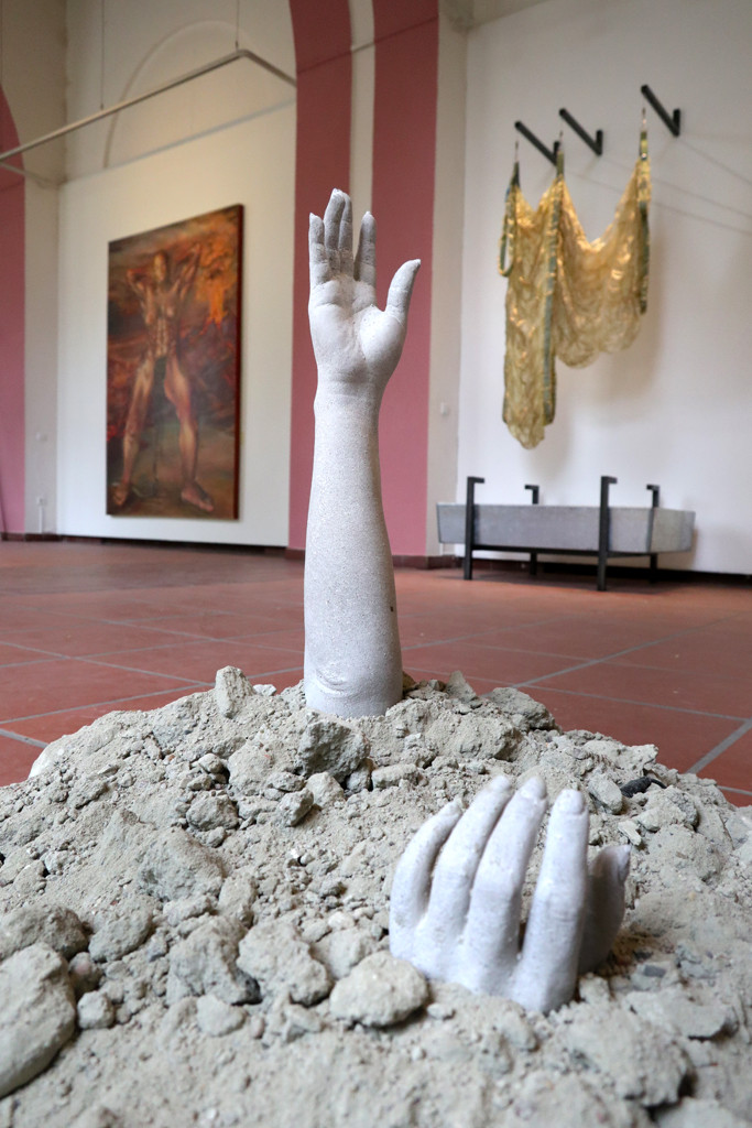 Aus einem Schutthaufen ragen ein Arm und eine Hand heraus, die aus Beton gegossen sind.