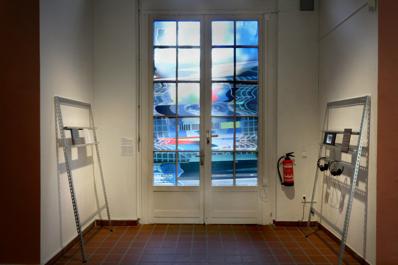 Ausstellungsansicht einer Mixed Media Installation. Mittig befindet sich ein mit bunter Fensterfolie beklebtes großes Fenster, rechts und links sind Metallhalterungen an die Wand gelehnt, auf denen Bücher, Kopfhörer, ein Video auf einem kleinen Bildschirm, und ein Text angebracht sind.