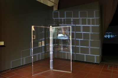Construction made of aluminum rods and transparent acrylic panels that mimic a glass revolving door. A video showing a wall-like structure consisting of rectangular blocks is projected onto the structure and the wall behind it.