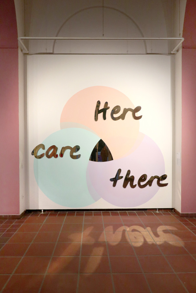 Drei an die Wand gemalte Farbkreise in Pastelltönen, die sich mittig überschneiden. In der sich überschneidenen Fläche ist ein Spiegel installiert. Über die Kreise sind in Spiegelacryl jeweils die Worte 