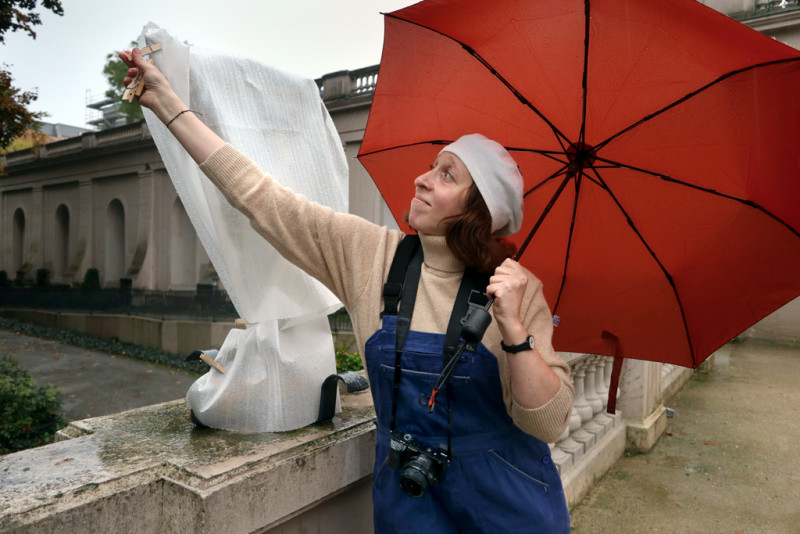 Man sieht eine Frau mit rotem Regenschirm die ein weiß verpacktes Kunstwerk entpackt
