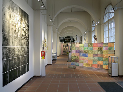 Links an der Wand ein Raster aus Bilder in Schwarz Weiß, in der Mitte farbige Drucke auf einem Bauzaum aufgehängt