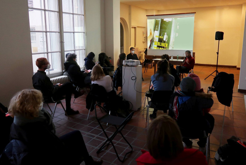 Lesung des Künstlers Omar Kasmani. Publikum sitzt auf Stühlen vor einer Leinwand auf der eine Präsentation abgespielt wird