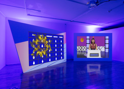Installationsansicht einer Kunstausstellung mit blau beleuchteten Fernsehstudio-Kulissen und einer Wettervorhersage-Präsentation