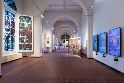 Langer Ausstellungsflur mit Rundbogendecke, seitlich farbenfrohe Kunstwerke in Vitrinen und an Wänden sowie digitale Bildschirme mit Ausstellungsinformationen