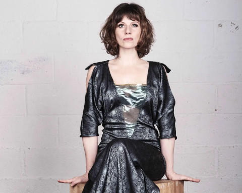 A woman sits on a wooden table wearing a grey gown.