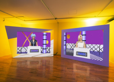 Kunstinstallation mit zwei großformatigen Bildern von Personen an Podien in einem gelben Ausstellungsraum