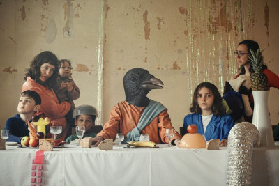 Surreale Tischszene mit Kindern und einer Figur mit Vogelkopf, die an einem gedeckten Tisch sitzen, vor einer verwitterten Wand